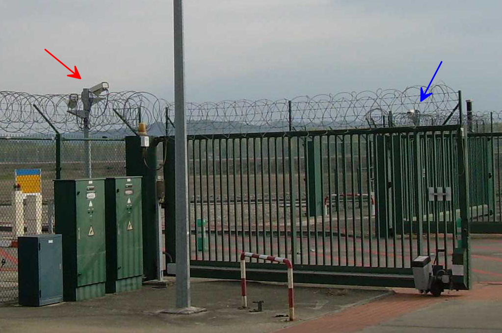 Des caméras à droite de la grille d'entrée de la centrale, et un objet muni d'une lumière à gauche.